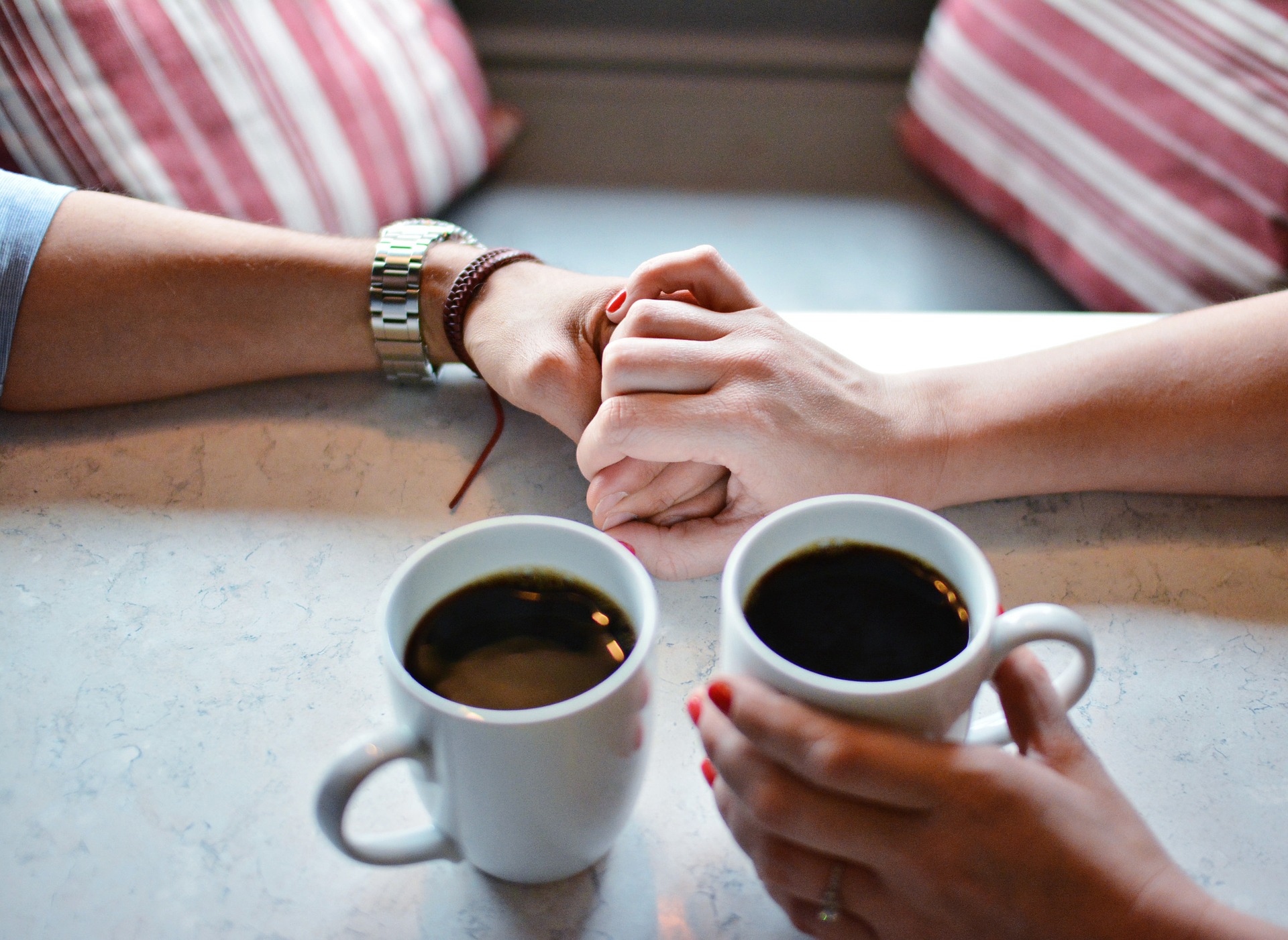 Närbild på två personers händer. Två koppar med kaffe i står på ett bord. Den ena personen håller den andras hand ömt tvärs över bordet.